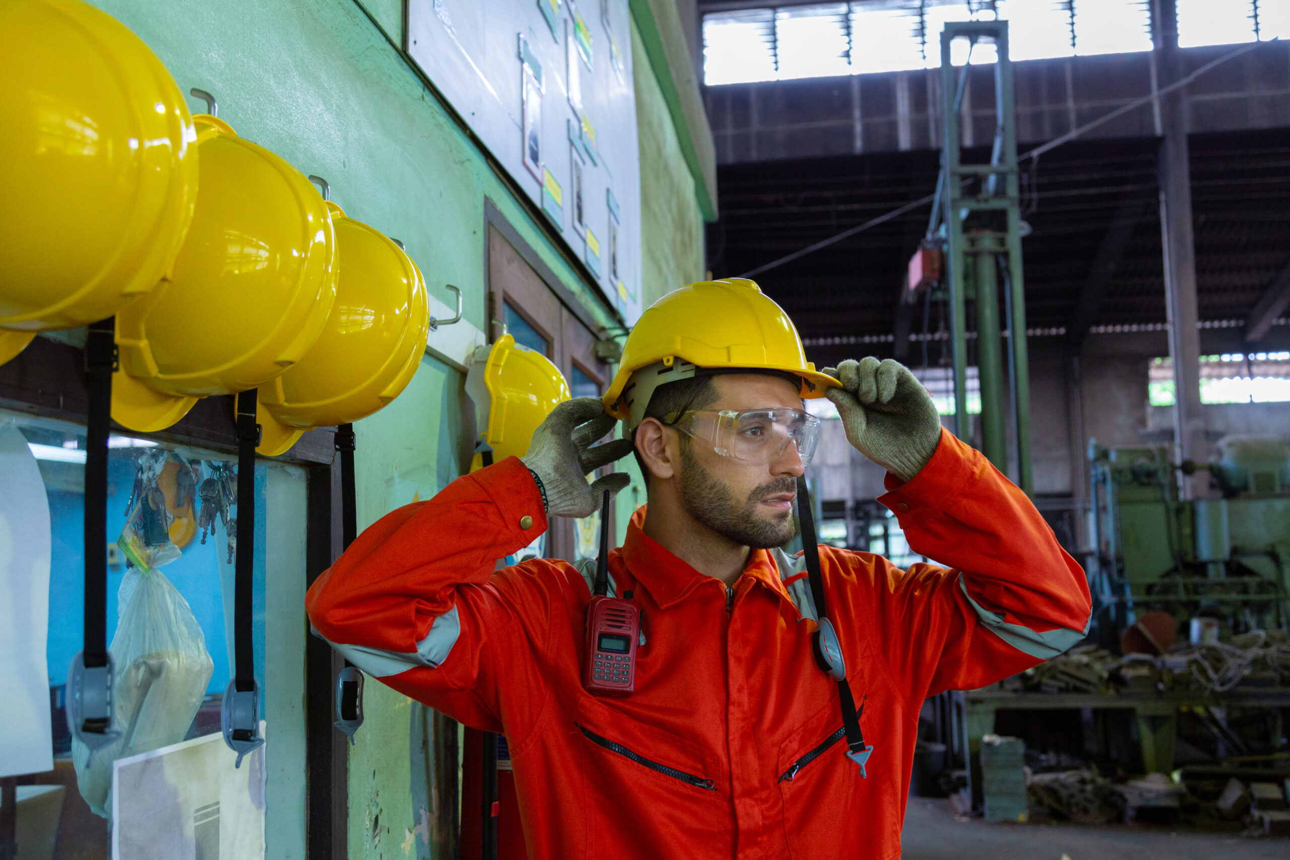 Trabajador industrial con casco amarillo y gafas de protección colocándose los EPIs en nave industrial. 
