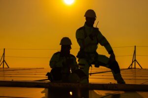 Dos trabajadores con EPIs y casco de seguridad instalando paneles solares bajo el sol en condiciones de calor extremo.